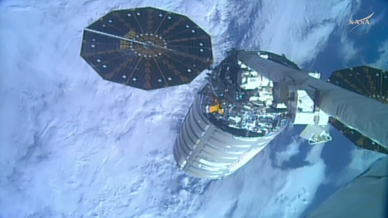 a cylindrical spacecraft at the end of a white mechanical arm above a blue and white planet