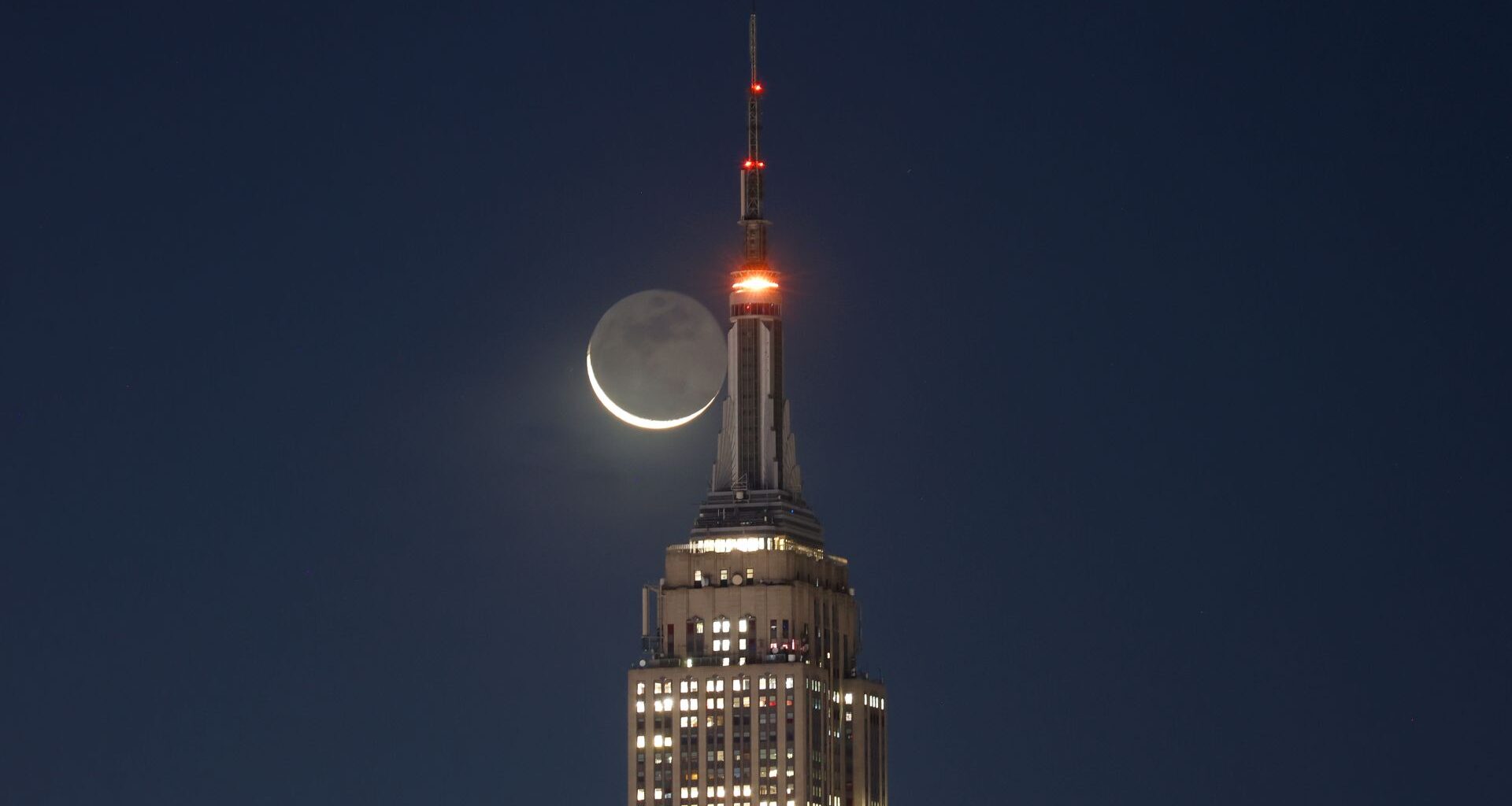 A thin crescent moon is shown next to the upper spire of a skyscraper in a dark sky. Its unlit surface is suffused with a soft glow, revealing the presence of lunar seas, or mare, on its surface.
