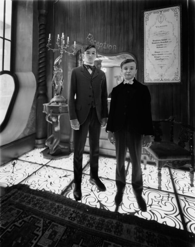 Two boys in vintage suits stand in a hotel reception area with ornate decor, a patterned floor, candelabra, statue, and framed sign on the wall. The scene has an old-fashioned, black-and-white aesthetic.
