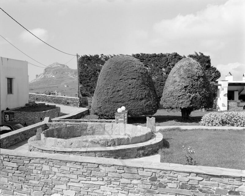 A black-and-white photo of a stone circular structure in a walled yard with neatly trimmed, dome-shaped trees and a building in the background. A mountain is visible in the distance under a partly cloudy sky.