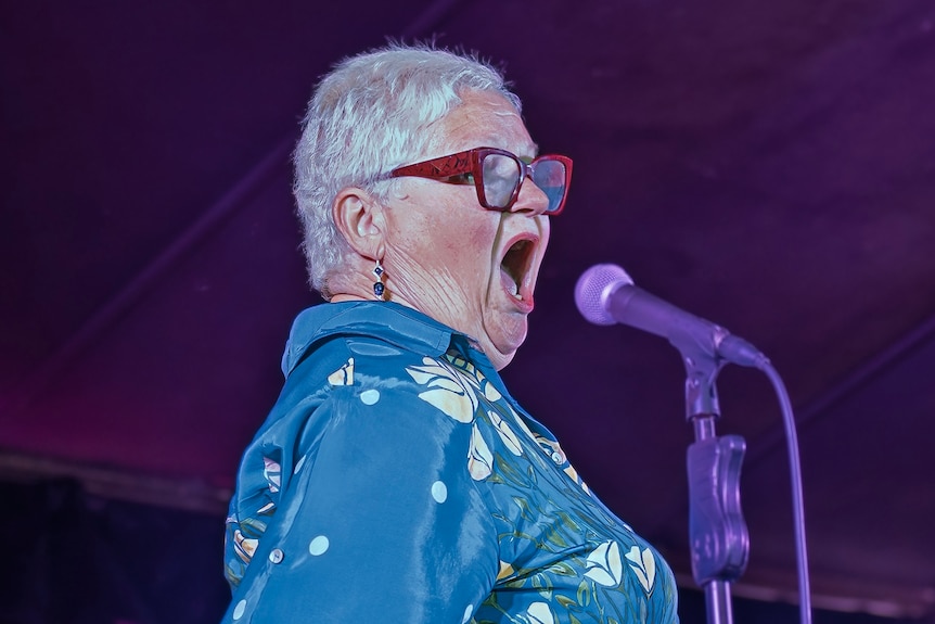 A grey haired woman in a blue outfit stands in front of a microphone.
