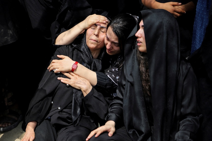 Three women dressed in black with black hijabs embrace each other as they grieve