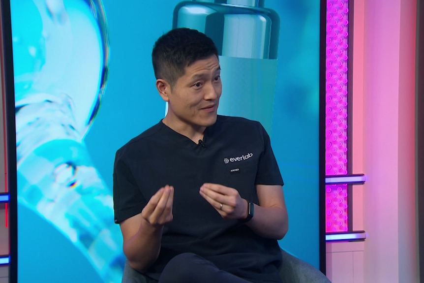Doctor wearing scrubs sitting and talking, in front of a screen with a generic image of a vial.