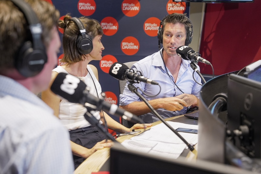 A man speaking into a microphone inside a radio studio, with two other people seated next to him.