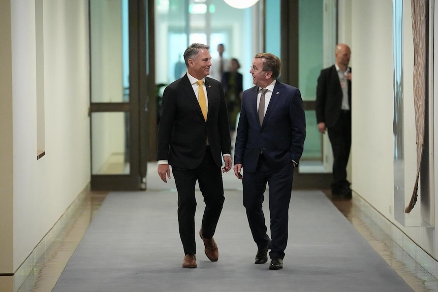 Two men in suits walk down a hallway.