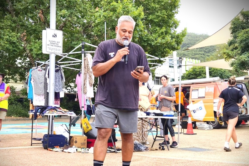 Man in a black tshirt and grey shorts speaks into a microphone, while in front of a second hand clothing rack.
