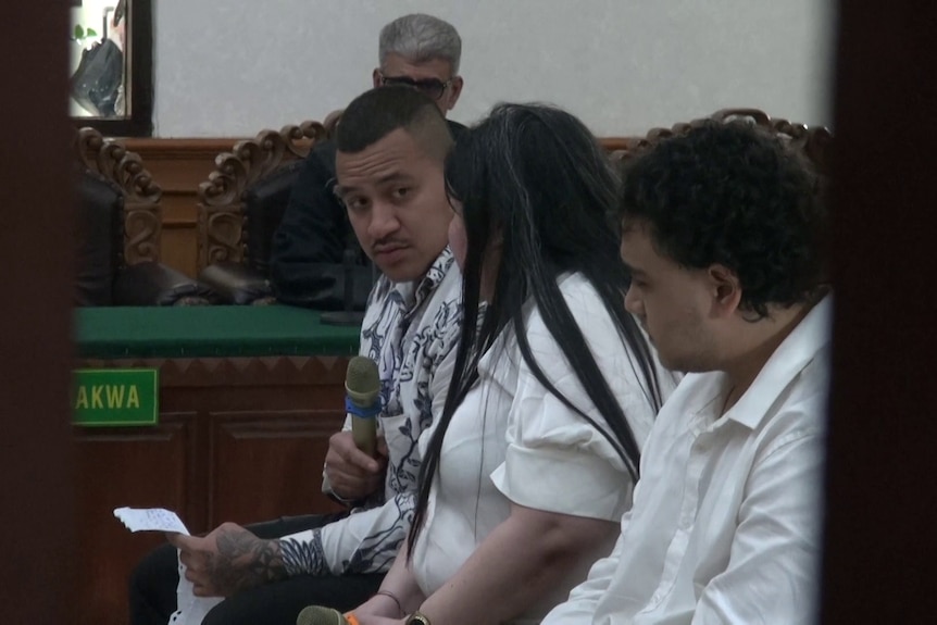 Two men in white collared shirts sit with a lawyer in court.