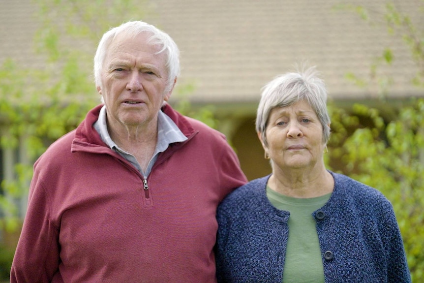 An older man in a red jumper standing next to a woman in a blue jumper and green shirt. Both have grey hair.