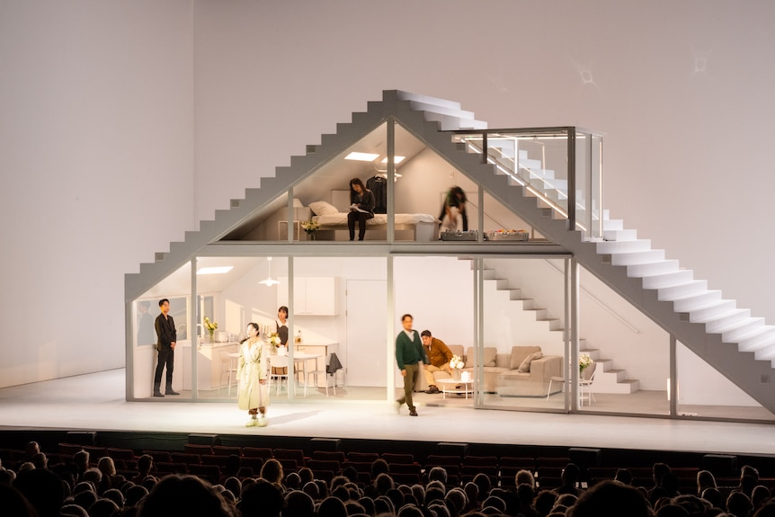 On stage is white house with floor-to-ceiling windows, and stairs on the outside, peaking at the roof. Korean actors are inside.