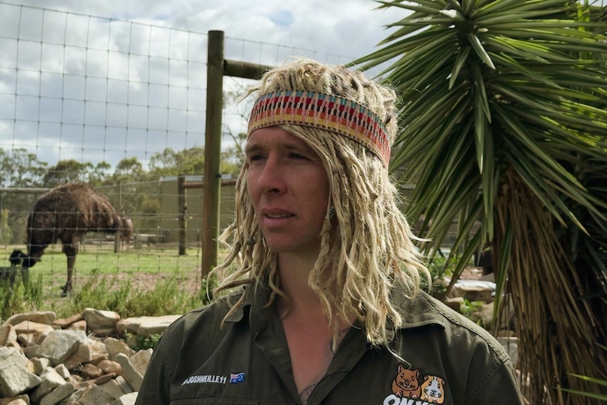 A photo of a man with blonde dreadlocks, wearing a green shirt, standing on a farm with emus in background.