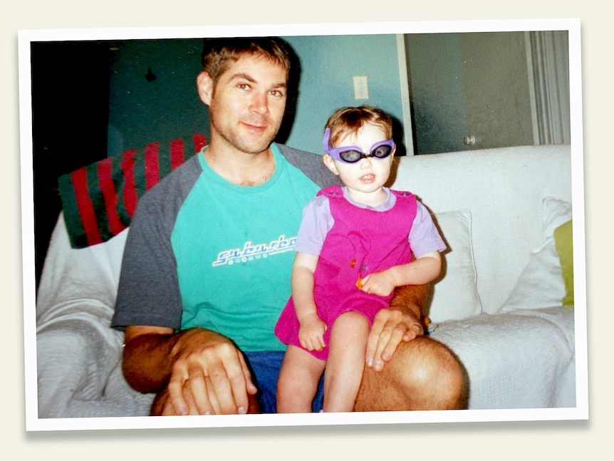 A man sits on a lounge and has a young girl wearing swimming goggles on his knee.