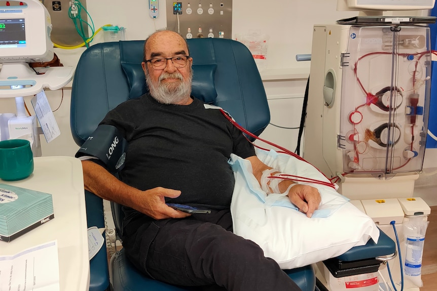 A 72 year-old man sits with several lines from machines leading to his arm and upper body.