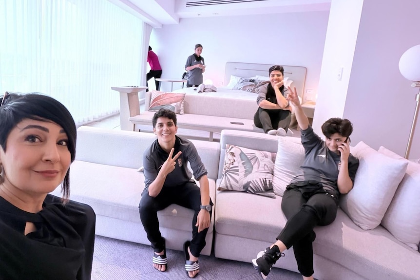A woman takes a selfie in a hotel room, with four other women in the background smiling