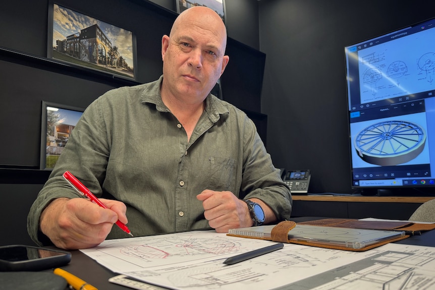 A man sits at a desk holding a red pen above drawings - screens with designs behind him. 
