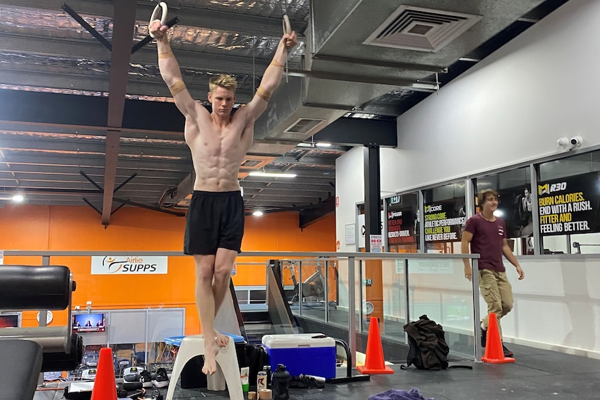 A young shirtless man hangs from two rings.