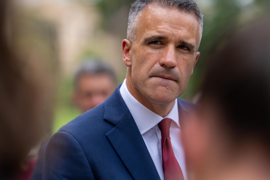 A man in a suit and red tie looks to the left of frame with a neutral expression.