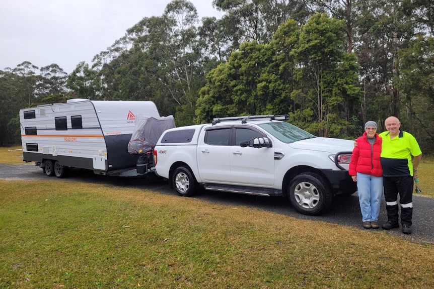 An retiree couple standing beside their white car and caravan. 