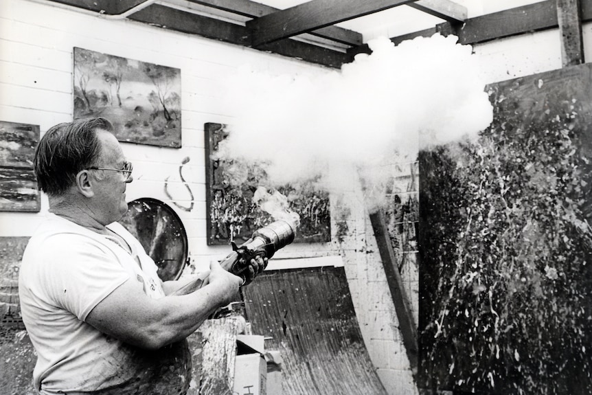 A black and white photo of a man holding a fired gun in front of a canvas splattered with paint. 