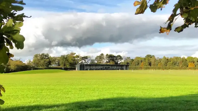 A large green field, with sun shining on the grass.  There is a long building made of a number of bays across the back of the field