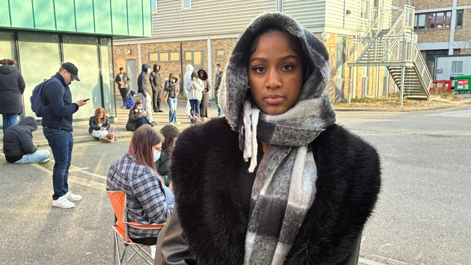 Young woman wearing hood, scarf and coat with others queuing outside behind her outdoors