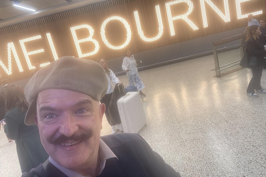 A smiling man with a moustache wears a beret while standing in an airport.
