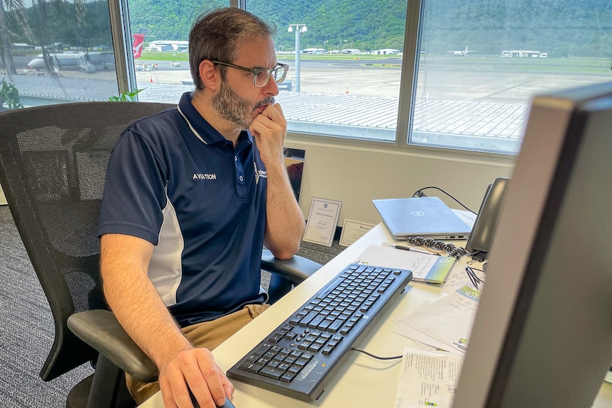 Man in blue polo looking at computer in office with airport in background through window