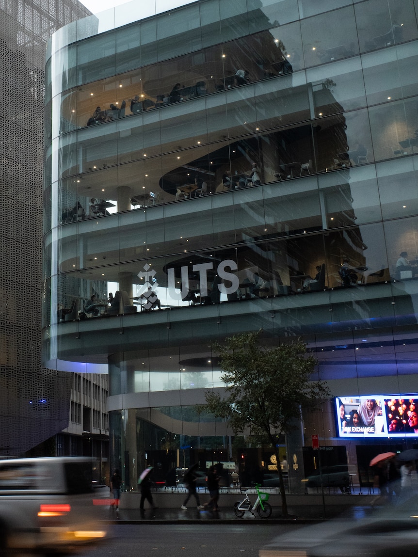 Traffic passes a modern glass-walled building at dusk. People can be seen inside at desks. Outside people have umbrellas.