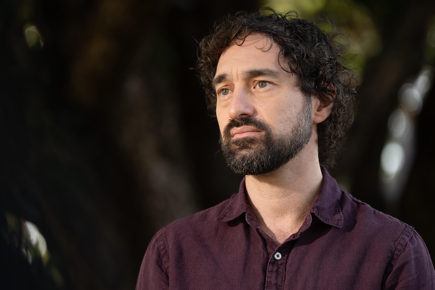 A man with dark curly hair and a beard wears a serious facial expression as he looks into the distance.