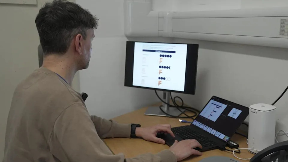 A man with short dark hair sat at a desk with a laptop and monitor. He is wearing a brown jumper. The room has white walls and the desk is made from light wood. There is a phone on the desk.