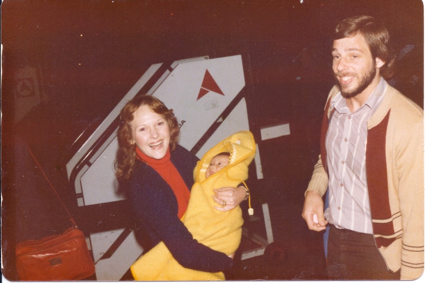 An old photo of a woman holding a baby wrapped in a yellow jacket and a man standing next to them 