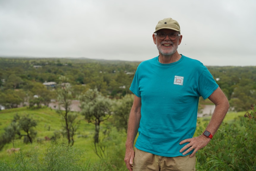 White man wearing blue t-shirt, beige cap and shorts, standing on greenside hills, hand on hip, round blue glasses.