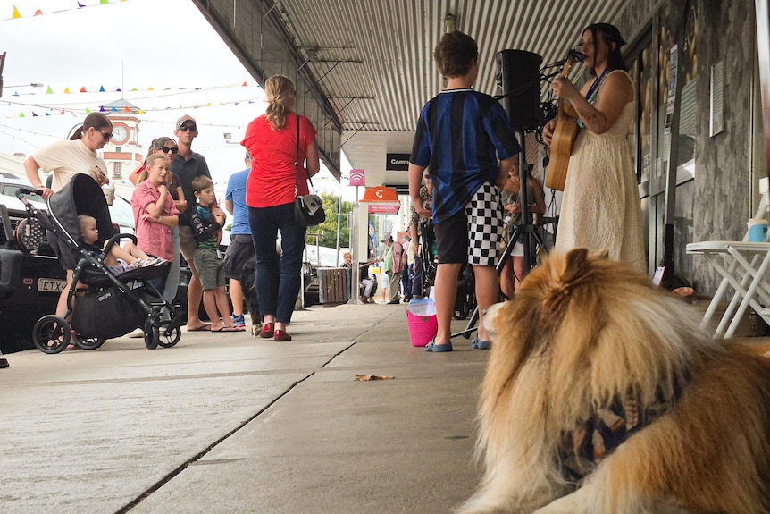 A country town street with performers, and a crowd. There is a dog in the foreground who also appears to be listening.