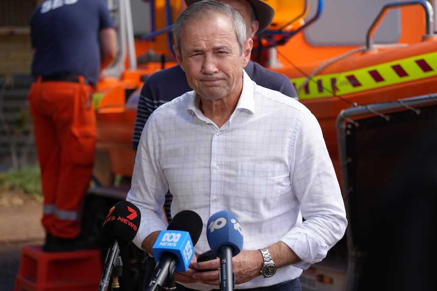A grey-haired man in a business shirt faces the media while standing in front of an emergency service truck.