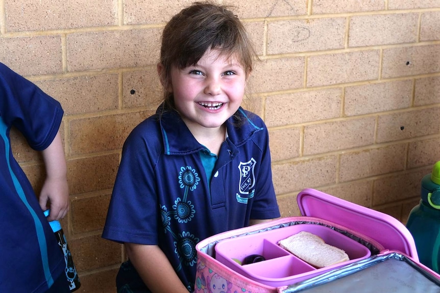 A young girl in a blue school uniform, sitting down with a pink lunchbox in front of her.