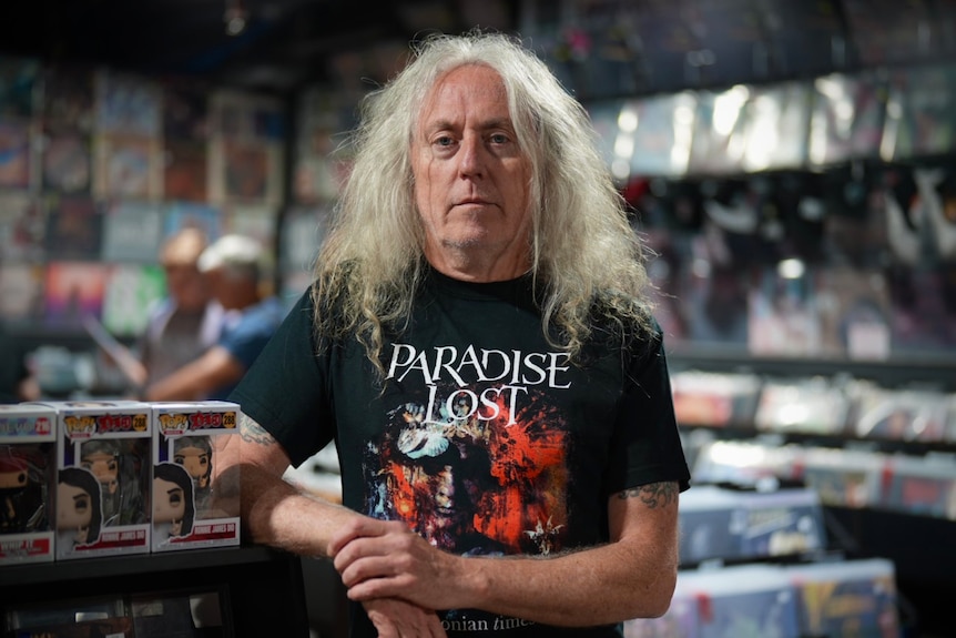 Mr Murphy with long white hair stands in a record shop wearing a Paradise Lost shirt with records behind him.
