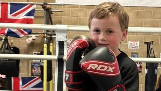 Jesse standing inside a boxing ring at a gym. He is wearing red and black gloves and holding them up in a fighting stance. He is smiling.
