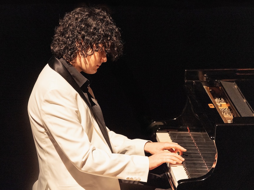 Malik sits at a piano with his hands on the keys, wearing a white suit jacket