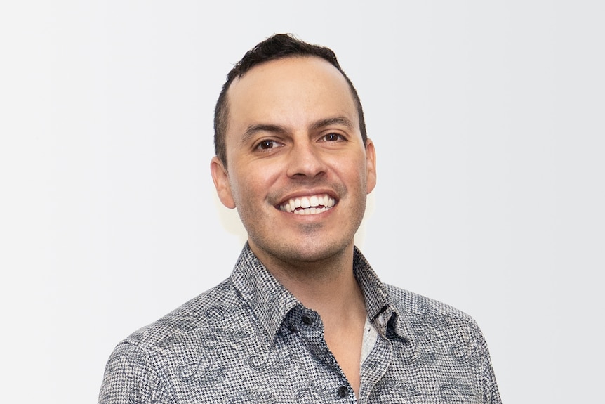 Man in grey collard shirt smiles at the camera in front of white background.
