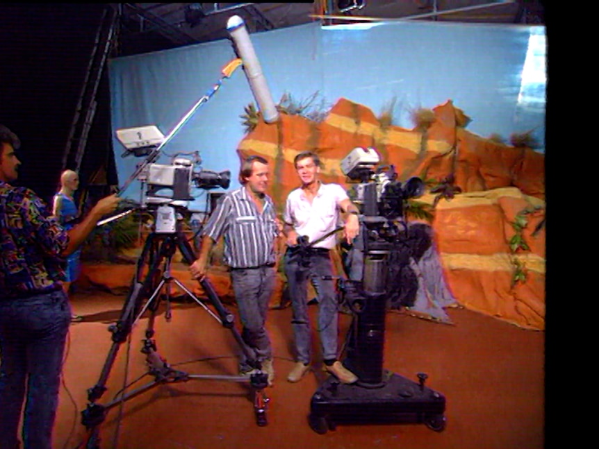 Two men in jeans and shirts stand in front of coloured background in a studio. A camera man with boom in front