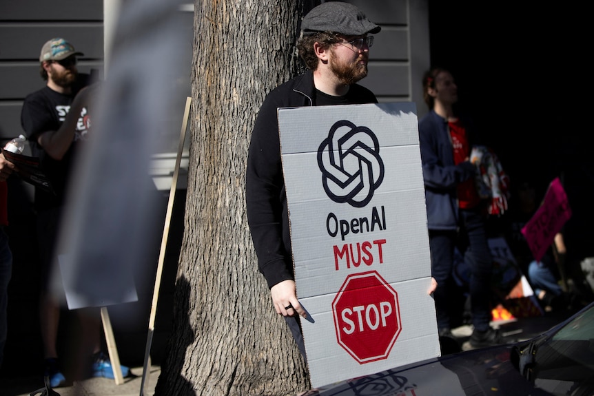 A man in a black top and grey cap holding a white, black and red 'OpenAI Must Stop' protest placard.