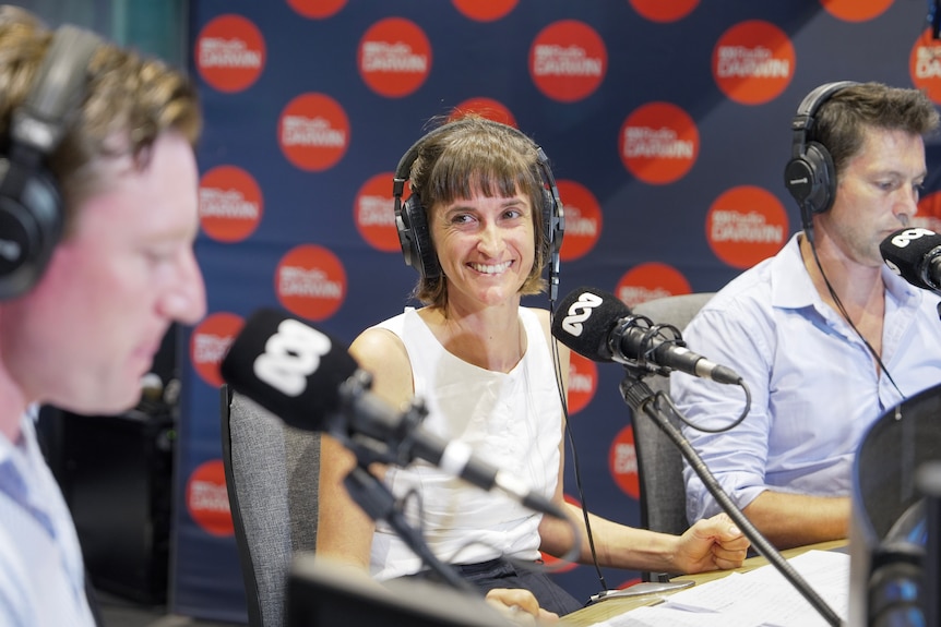 A woman, smiling widely, sitting in front of a microphone inside a radio studio, flanked by two men.