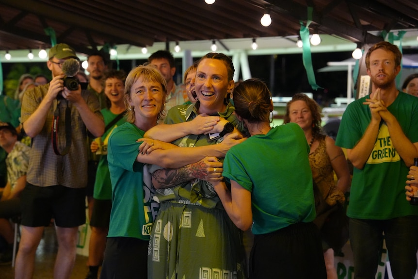 A person, white, wearing green shirt and pants, smiling, surrounded by people wearing green 'The Greens' shirts.