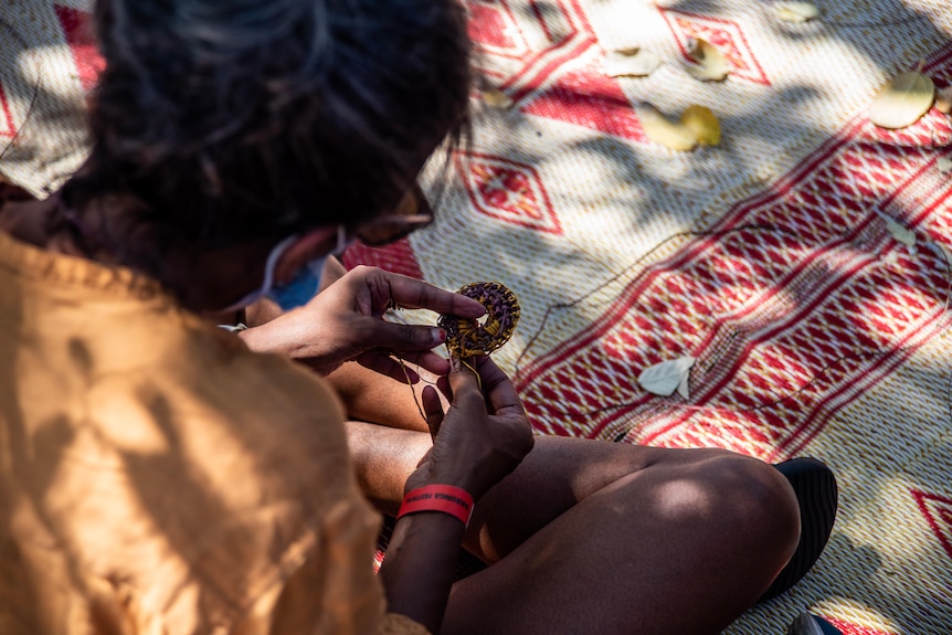 Photo taken from behind of an Indigenous woman sitting cross-legged an holds something in hand, sits on white and red rug.