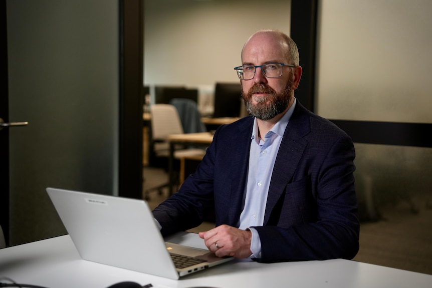Man wearing glasses looks at camera from behind his laptop.