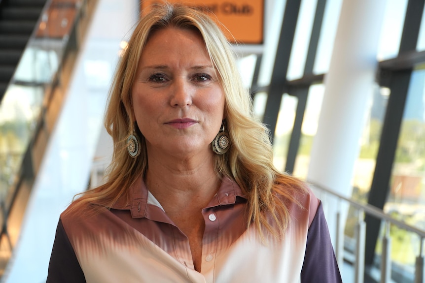 A blonde woman stands looking seriously at the camera. 