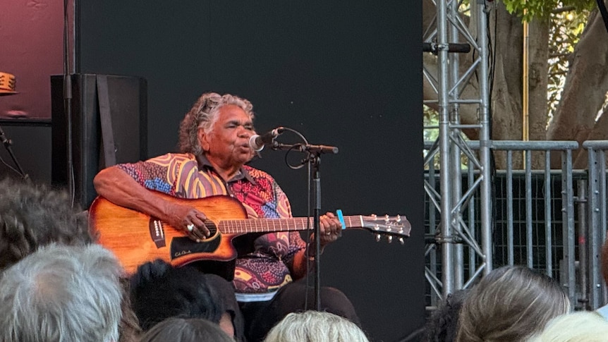 A performer plays the guitar on an outdoor festival stage with a crowd watching in front.