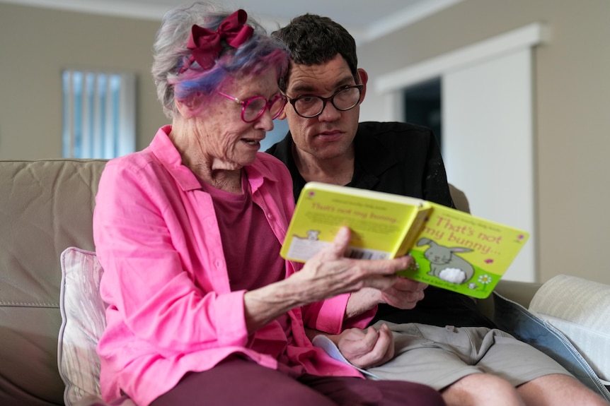 A woman in all pink from her outfit to her glasses and hair highlights holds a picture book while her adult son sits beside
