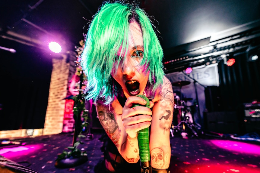 A woman with a green messy bob and tattoos sings into a green mic under purple lights on a stage.