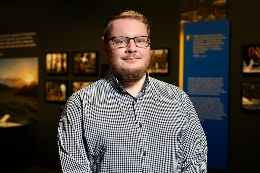 A man with red hair stands in front of a wall at a museum smiling