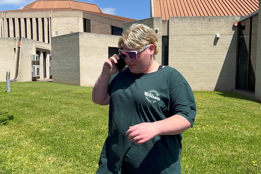 A young woman with short blonde hair wearing sunglasses and a T-shirt outside a concrete building